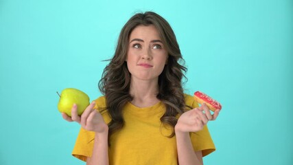 Poster - A beautiful young woman wearing a yellow t-shirt is trying to choose between apple and donut standing isolated over blue background