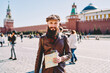 © BullRun - Half length portrait of smiling man traveller holding map for navigation and looking at camera with Red Square on background, positive hipster guy dressed in trendy wear exploring Moscow city