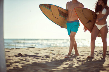 Wall Mural - cropped image of young fit sexy caucasian couple walking on sandy beach holding surfboards, copy space