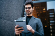 © BullRun - Below view of successful male entrepreneur dressed in elegant apparel downloading file with last business news on modern touch pad device connected to 4G internet standing near office building