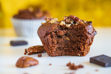 Healthy muffin with a bite from the center a homemade freshly baked and gluten free with pecans and cocoa powder and chocolate cubes on white table with yellow blurred background, nutrition concept