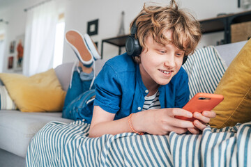 Preteen boy lying at home on cozy sofa dressed casual jeans and shirt. listening to music and chatting using wireless headphones connected with smartphone. Child using electronic devices concept