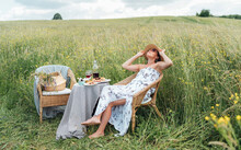 Young Woman Dressed Light Summer Dress And Straw Hat Sitting In A Rattan Chair On The High Green Grass Meadow And Enjoying The Moment Near The Picnic Table With Red Wine And Homemade Pie.