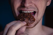 © Kate - A detailed close-up shot of the mouth of a young handsome man who bites a cookie, holding it in his hand on a home background. Man eating chocolate cookie with peanut, white teeth, close up face.