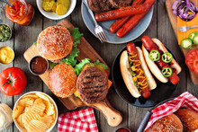 Summer BBQ Food Table Scene With Hot Dog And Hamburger Buffet. Top View Over A Dark Wood Background.