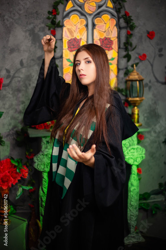 Cuadros en Lienzo Young woman in a black cloak with a magic wand in her hands, standing against the backdrop of a medieval window