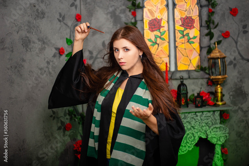 Fotomural Young woman in a black cloak with a magic wand in her hands, standing against the backdrop of a medieval window