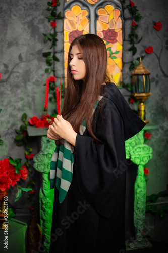 Fotografía Young woman in a black cloak with a magic wand in her hands, standing against the backdrop of a medieval window