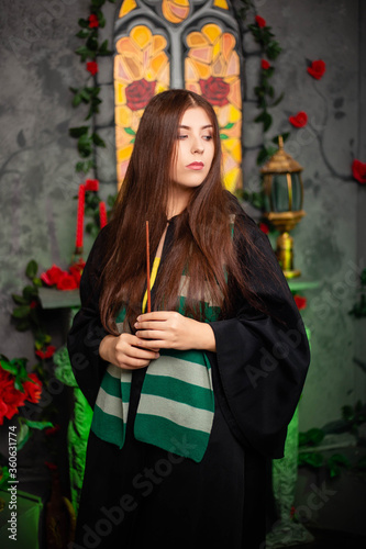 Fotomural Young woman in a black cloak with a magic wand in her hands, standing against the backdrop of a medieval window