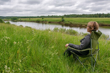 Wall Mural - Young woman sitting in the outdoor chair on the bank of Ugra river. Ugra national park, Kaluga oblast, Russia.