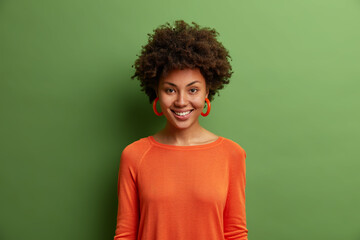 Canvas Print - Portrait of beautiful dark skinned woman with bushy curly hair, smiles toothily, has optimistic look, wears orange jumper and earrings looks directly at camera glad to hear good news isolated on green