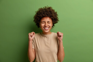 Lucky positive woman with Afro hair clenches fists with triumph, celebrates dream come true, smiles broadly with closed eyes, enjoys achievement, becomes champion, isolated on green background