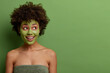 © wayhome.studio  - Lovely happy woman with curly hair, looks aside smiling, wears green beauty mask for skin pampering, cares about complexion, stands wrapped in bath towel, bare shoulders, isolated on green background