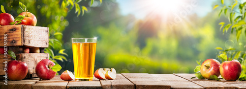 Fotografía Apple Crate And Glass Of Juice On Wooden Table With Sunny Orchard Background - A