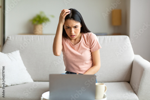 Cuadros en Lienzo Puzzled Girl At Laptop Scratching Head Sitting On Couch Indoor