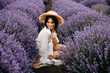 Young beautiful woman sitting in lavender field, wearing a bohemian white dress and a straw hat.