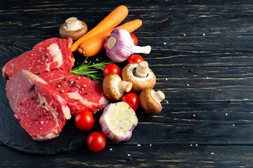 Wall Mural - Three pieces of juicy raw beef with vegetables on a stone cutting board on a black wooden table background.