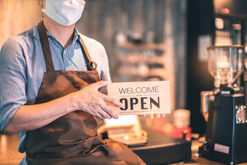 Business owner attractive young Asian man in apron hanging we're open sign on front door  welcoming clients to new cafe. Happy waiter with protective face mask holding open sign.