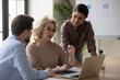 © fizkes - Diverse employees listening to mature mentor coach training staff interns, pointing at laptop screen, middle aged businesswoman wearing glasses with colleagues working on project together