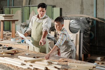 Wall Mural - Skilled carpenter in apron pointing at wooden detail while teaching son to make furniture in workshop