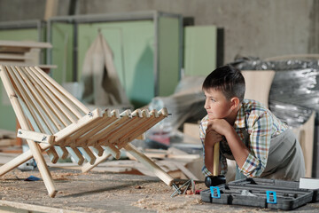 Wall Mural - Pensive teenage carpenter leaning on hammer while looking at his finished wooden chair in workshop