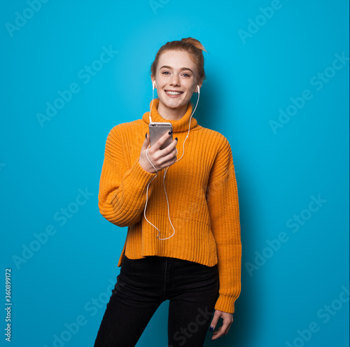 Caucasian ginger woman with freckles smiling at camera while listening to music and holding a phone on blue studio wall