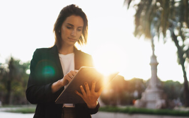 Wall Mural - Successful female banker using tablet and wireless earphones outdoor background sun lights, portrait young woman professional manager working on touch pad in evening city