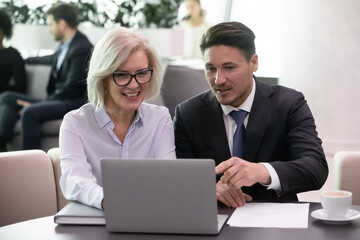 Business man and 50 years old woman colleagues looking at laptop happy smiling. Businessman and businesswoman sitting and working together discussing successful strategy results on computer monitor