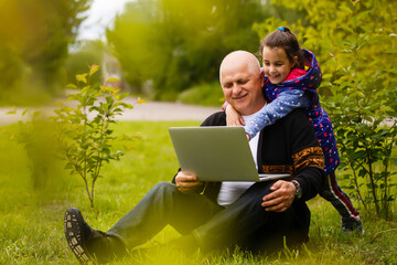 Senior man with his granddaughter is studying a laptop outdoors. Internet education for old grandfather.