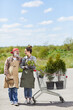 © Seventyfour - Vertical full length portrait of two gardeners holding flower pots looking at each other while standing by cart outdoors, copy space
