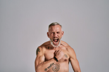 Unstoppable. Half naked middle aged muscular man shouting at camera, beating his chest while posing in studio over grey background