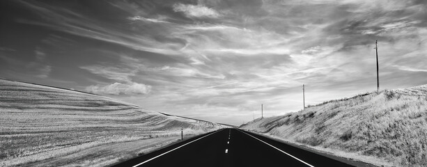 Black and white photo of 2 lane highway next to harvested wheat fields heading into distance 