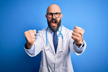 Wall Mural - Handsome bald doctor man with beard wearing glasses and stethoscope over blue background angry and mad raising fists frustrated and furious while shouting with anger. Rage and aggressive concept.