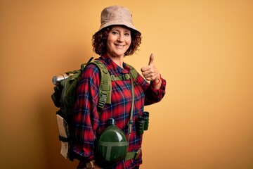 Poster - Middle age curly hair hiker woman hiking wearing backpack and water canteen using binoculars doing happy thumbs up gesture with hand. Approving expression looking at the camera showing success.