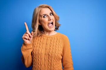 Middle age beautiful blonde woman wearing casual sweater over yellow background pointing finger up with successful idea. Exited and happy. Number one.