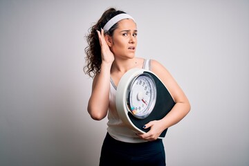 Sticker - Young beautiful woman with curly hair holding weighing machine over white background smiling with hand over ear listening an hearing to rumor or gossip. Deafness concept.