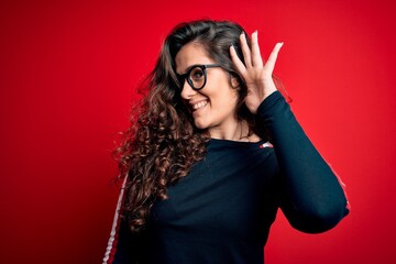 Canvas Print - Young beautiful woman with curly hair wearing sweater and glasses over red background smiling with hand over ear listening an hearing to rumor or gossip. Deafness concept.