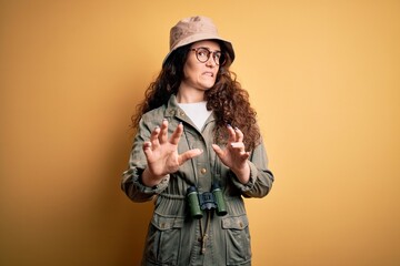 Poster - Young beautiful tourist woman on vacation wearing explorer hat and binoculars disgusted expression, displeased and fearful doing disgust face because aversion reaction. With hands raised