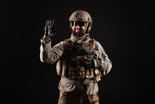 USA Soldier In A Military Suit With A Rifle Smiles And Shows The Okay Sign Against A Dark Background, An American Commando In Uniform