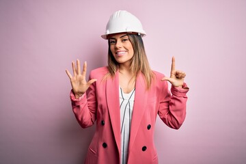 Poster - Young beautiful brunette architect woman wearing safety helmet over pink background showing and pointing up with fingers number seven while smiling confident and happy.