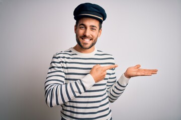 Wall Mural - Young handsome sailor man with beard wearing navy striped uniform and captain hat amazed and smiling to the camera while presenting with hand and pointing with finger.