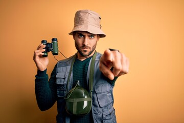 Canvas Print - Young handsome tourist man with beard on vacation wearing explorer hat using binoculars pointing with finger to the camera and to you, hand sign, positive and confident gesture from the front