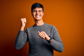 Sticker - Young handsome man wearing casual sweater standing over isolated orange background Pointing to the back behind with hand and thumbs up, smiling confident