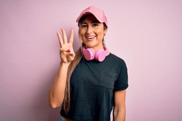 Wall Mural - Young beautiful fitness sports woman wearing training cap and headphones over pink background showing and pointing up with fingers number three while smiling confident and happy.