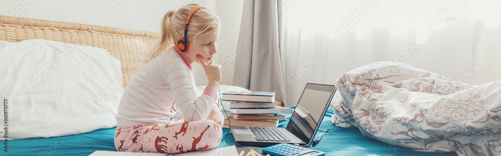 Girl child sitting in bed and learning online on laptop Internet. Virtual class lesson on video during self isolation quarantine. Distant remote education class. Web banner header for a website. - obrazy, fototapety, plakaty 