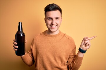 Wall Mural - Young handsome caucasian man drinking a bottle of beer over yellow background very happy pointing with hand and finger to the side