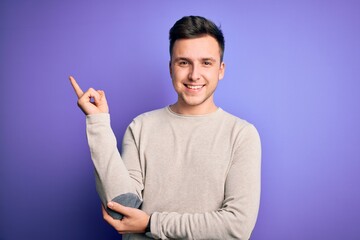 Wall Mural - Young handsome caucasian man wearing casual sweater over purple isolated background with a big smile on face, pointing with hand and finger to the side looking at the camera.