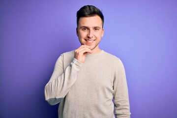 Wall Mural - Young handsome caucasian man wearing casual sweater over purple isolated background looking confident at the camera with smile with crossed arms and hand raised on chin. Thinking positive.