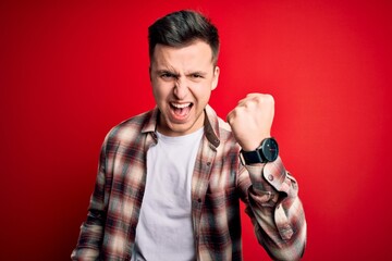 Wall Mural - Young handsome caucasian man wearing casual modern shirt over red isolated background angry and mad raising fist frustrated and furious while shouting with anger. Rage and aggressive concept.