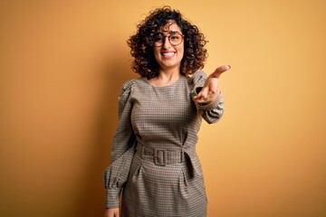 Sticker - Beautiful arab business woman wearing dress and glasses standing over yellow background smiling friendly offering handshake as greeting and welcoming. Successful business.